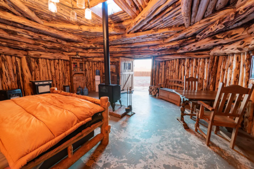 Cozy interior of a rustic cabin with wooden walls, a bed, dining table, and a wood stove. Natural light from a skylight.